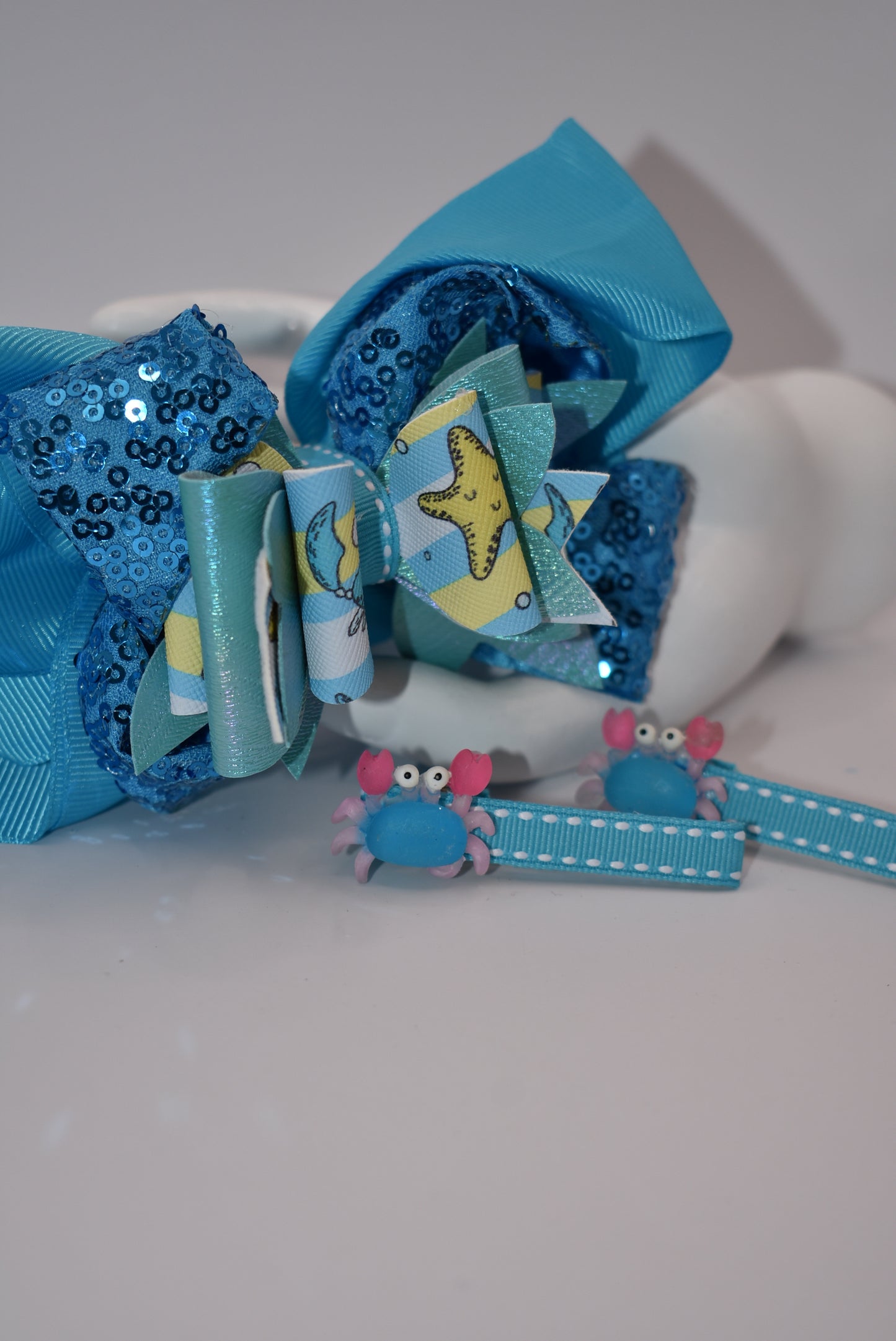 Get ready to make a splash with Nana's Collection! Our Under the Sea Bow features a playful crab-inspired design mixed with gorgeous blue and sparkle ribbons. This 3-piece set also includes two matching alligator clips for a complete look.