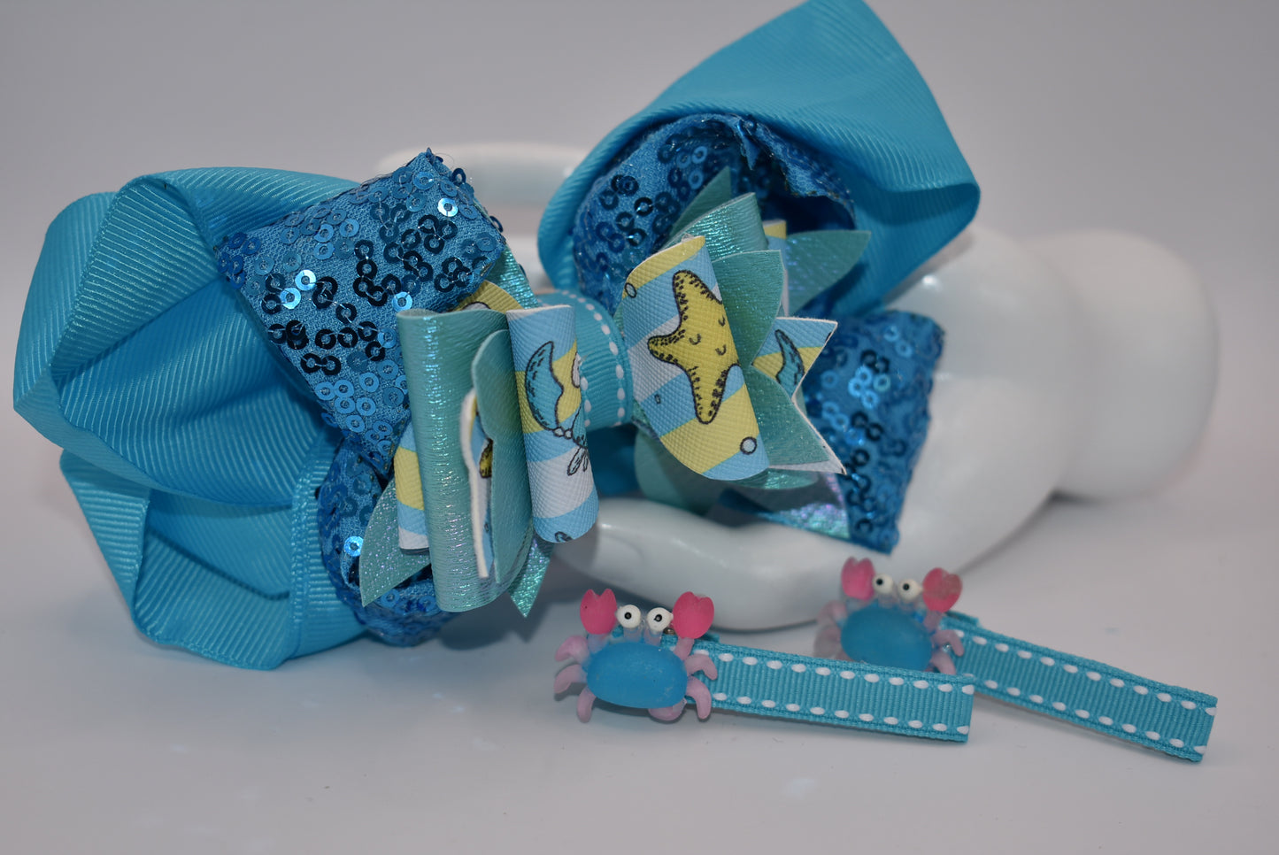 Get ready to make a splash with Nana's Collection! Our Under the Sea Bow features a playful crab-inspired design mixed with gorgeous blue and sparkle ribbons. This 3-piece set also includes two matching alligator clips for a complete look.