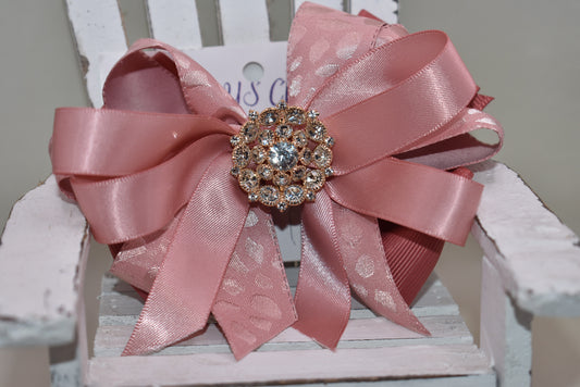 Add a touch of sparkle with this charming bow from Nana's Collection 180. Featuring a soft rose gold pink ribbon topped with a dazzling crystal charm, it’s the perfect playful accent for any outfit. The wrapped alligator clip ensures a secure fit all day long, so you can shine and twirl without a care. Ideal for dressing up your look anytime you want to add a bit of magic!