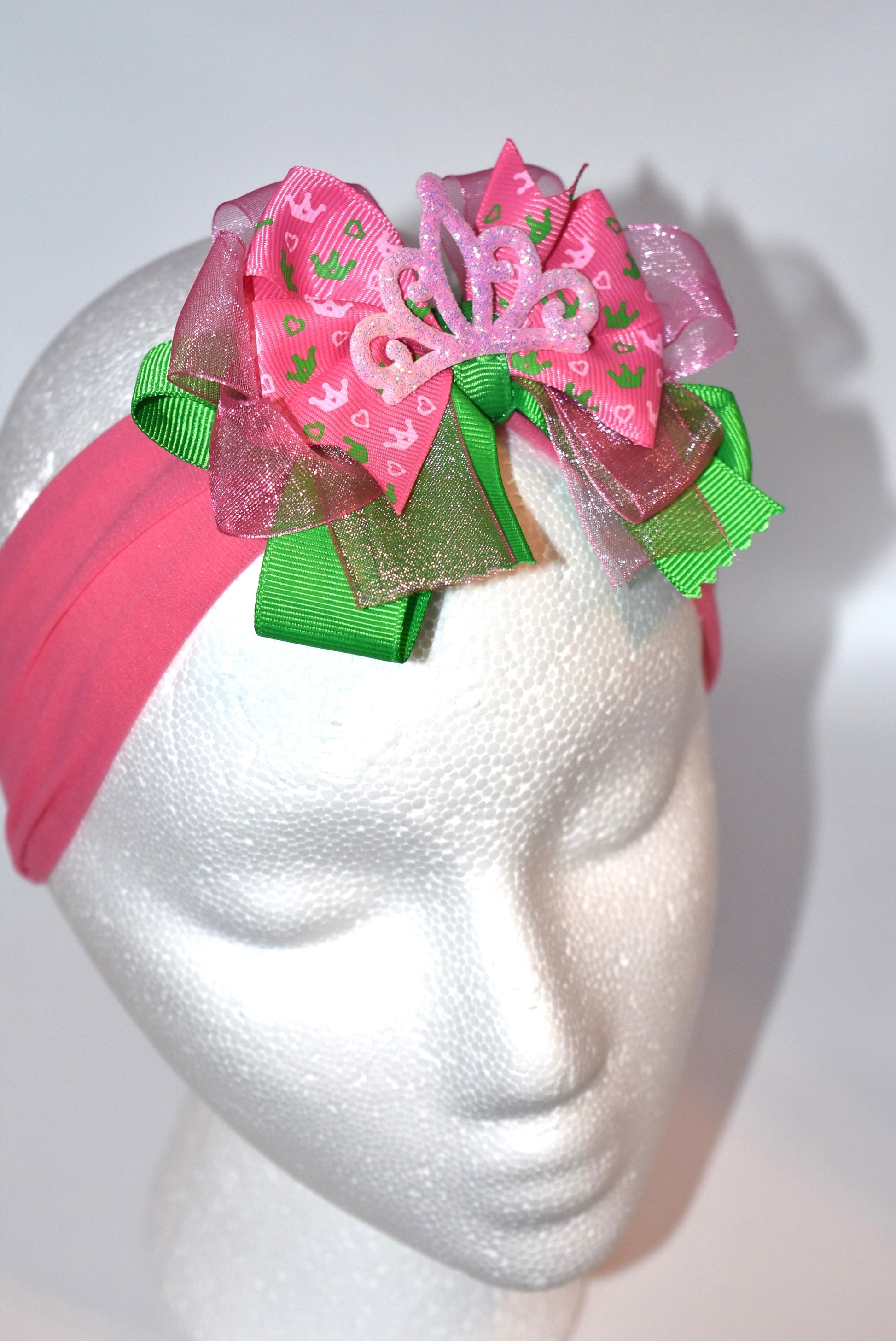 This one-of-a-kind wide headband features soft pink nylon for all-day comfort. Its adorable pink and green ribbon is topped with a playful princess bow and crown, adding a magical touch. Perfect for anyone young or old who loves a fun, charming accessory.
