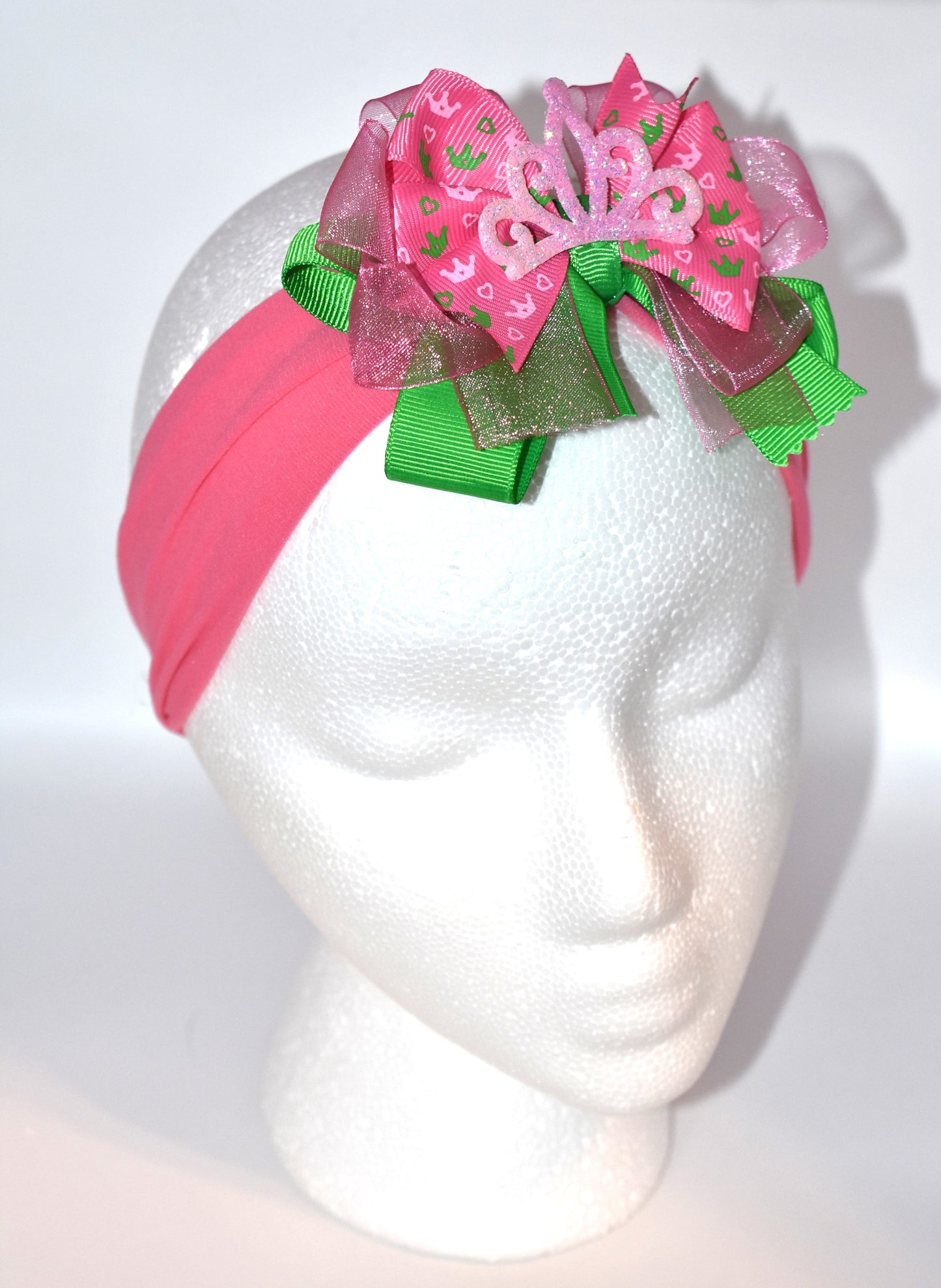 This one-of-a-kind wide headband features soft pink nylon for all-day comfort. Its adorable pink and green ribbon is topped with a playful princess bow and crown, adding a magical touch. Perfect for anyone young or old who loves a fun, charming accessory.