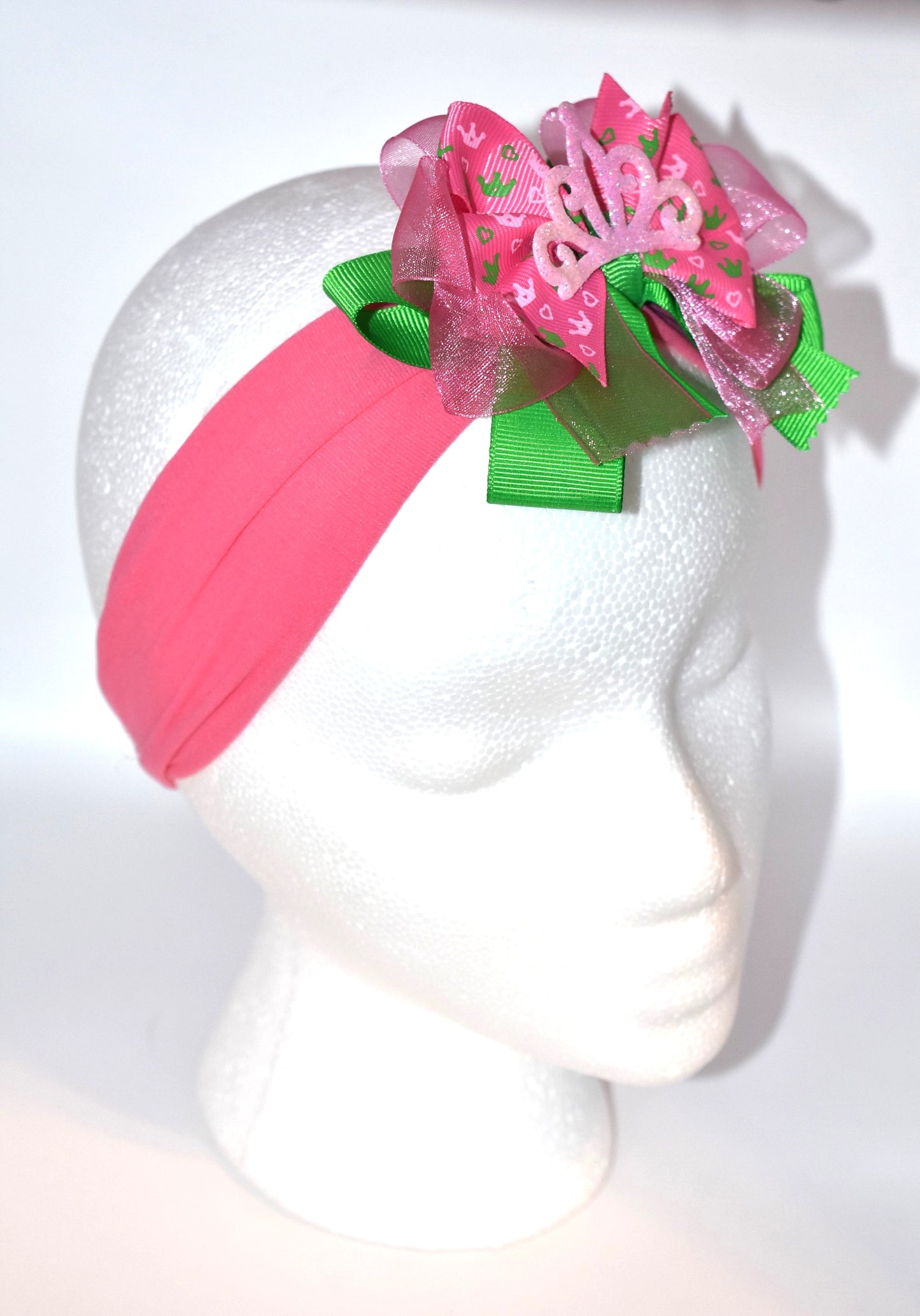 This one-of-a-kind wide headband features soft pink nylon for all-day comfort. Its adorable pink and green ribbon is topped with a playful princess bow and crown, adding a magical touch. Perfect for anyone young or old who loves a fun, charming accessory.