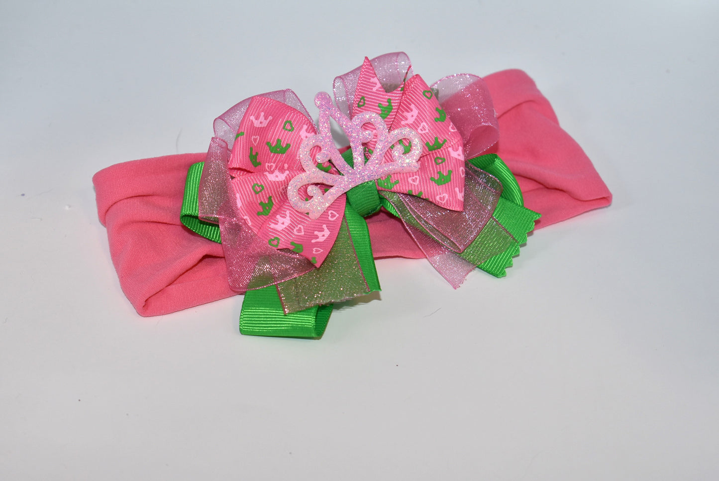 This one-of-a-kind wide headband features soft pink nylon for all-day comfort. Its adorable pink and green ribbon is topped with a playful princess bow and crown, adding a magical touch. Perfect for anyone young or old who loves a fun, charming accessory.