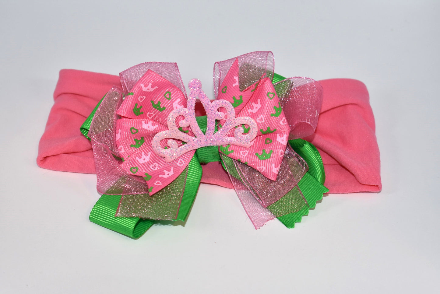 This one-of-a-kind wide headband features soft pink nylon for all-day comfort. Its adorable pink and green ribbon is topped with a playful princess bow and crown, adding a magical touch. Perfect for anyone young or old who loves a fun, charming accessory.