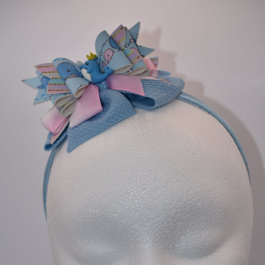 Introducing Nana's Collection, a one-of-a-kind piece featuring a playful whale motif, combined with a pretty blue and pink faux leather and ribbon design. Accompanied by a satin baby blue headband, this accessory is not only adorable but also super flexible and comfortable for all-day wear.