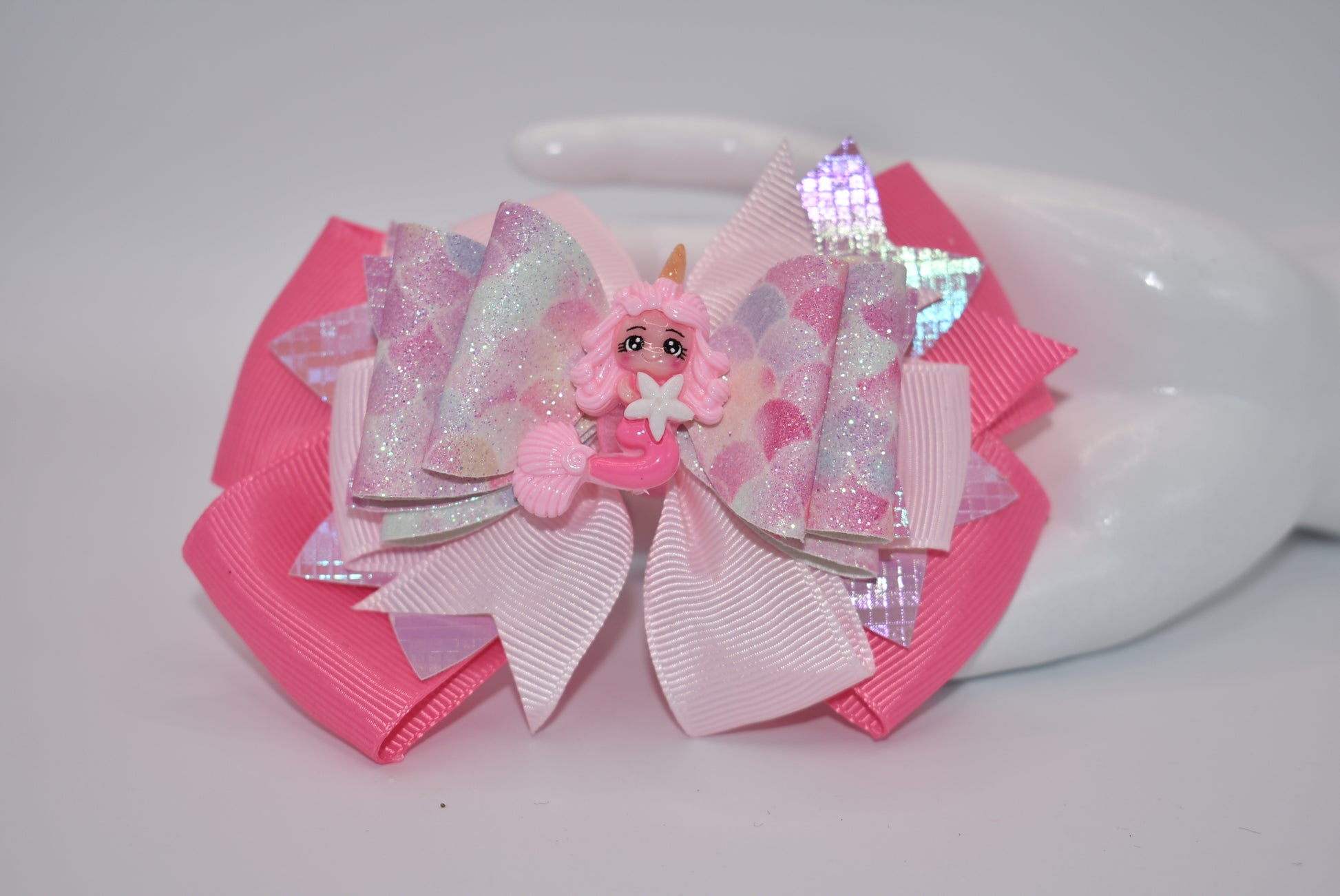 Add a touch of whimsy and sparkle to any outfit with Nana's Collection. This textured faux leather bow, in the prettiest shade of pink, features a playful mermaid design and layers of fun. The alligator clip ensures comfortable wear, while the pink ribbon bow adds a perfect finishing touch.