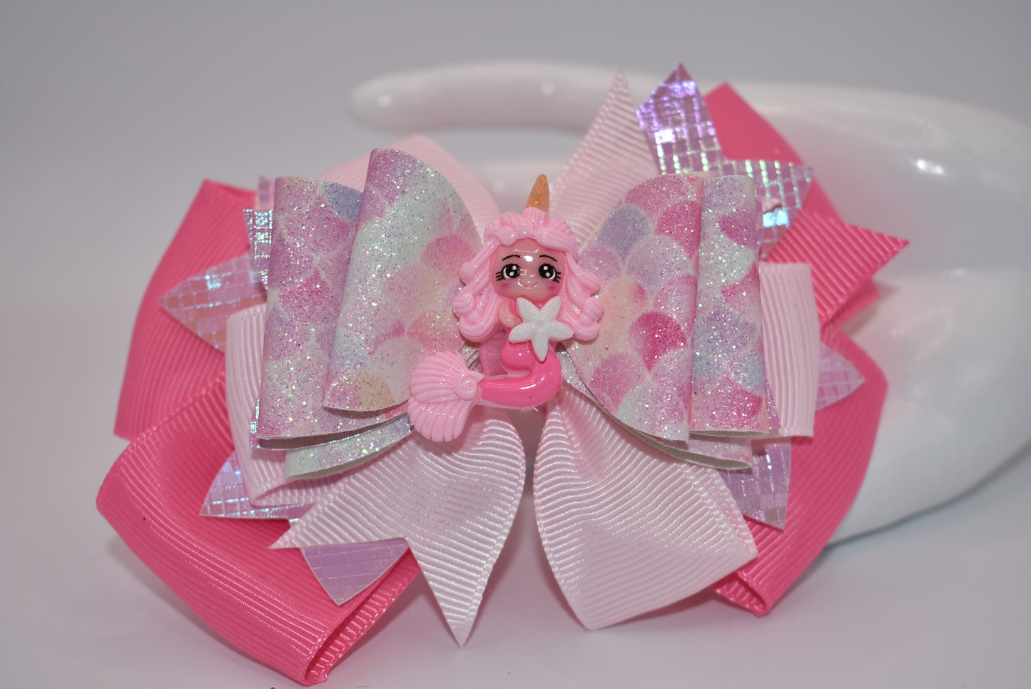 Add a touch of whimsy and sparkle to any outfit with Nana's Collection. This textured faux leather bow, in the prettiest shade of pink, features a playful mermaid design and layers of fun. The alligator clip ensures comfortable wear, while the pink ribbon bow adds a perfect finishing touch.