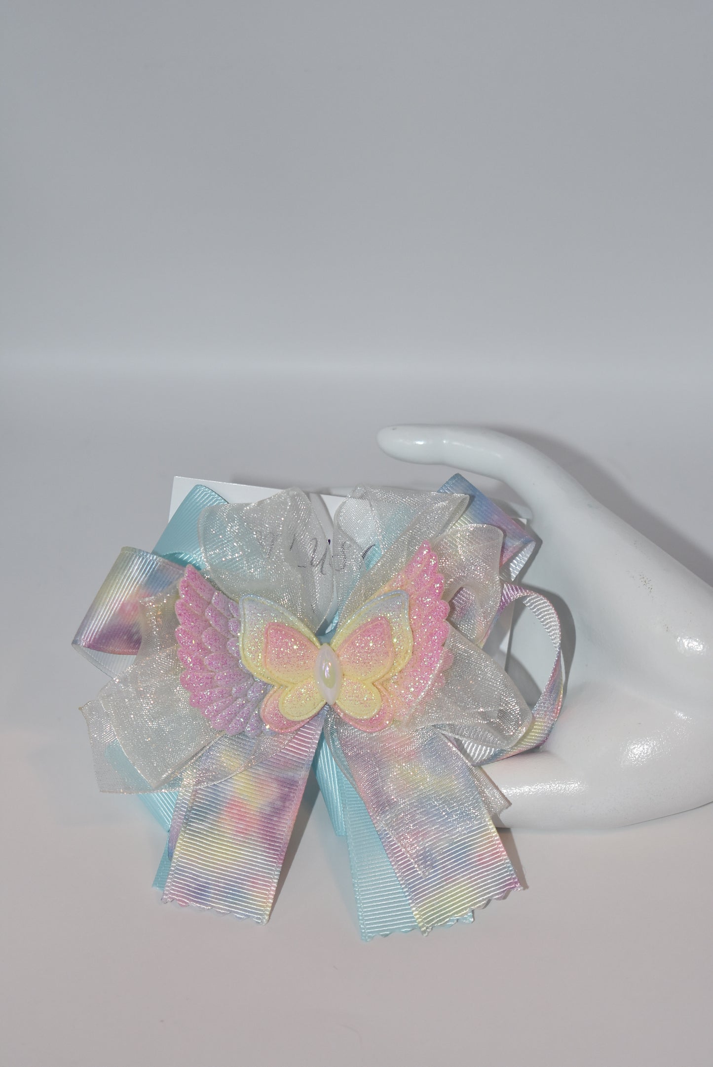 Add a splash of joy with this playful rainbow tie-dye ribbon featuring cheerful pink, blue, and yellow hues. A charming winged butterfly perches atop, bringing whimsy to your look. Securely attached to a wrapped alligator clip, it’s perfect for brightening any outfit with a touch of fun and color.