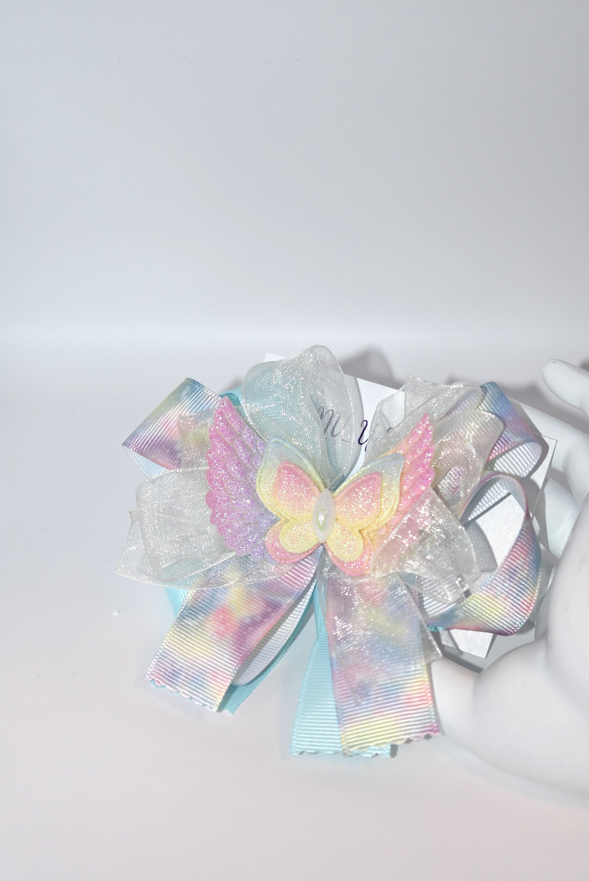Add a splash of joy with this playful rainbow tie-dye ribbon featuring cheerful pink, blue, and yellow hues. A charming winged butterfly perches atop, bringing whimsy to your look. Securely attached to a wrapped alligator clip, it’s perfect for brightening any outfit with a touch of fun and color.