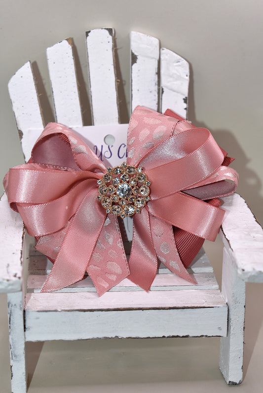 Add a touch of sparkle with this charming bow from Nana's Collection 180. Featuring a soft rose gold pink ribbon topped with a dazzling crystal charm, it’s the perfect playful accent for any outfit. The wrapped alligator clip ensures a secure fit all day long, so you can shine and twirl without a care. Ideal for dressing up your look anytime you want to add a bit of magic!
