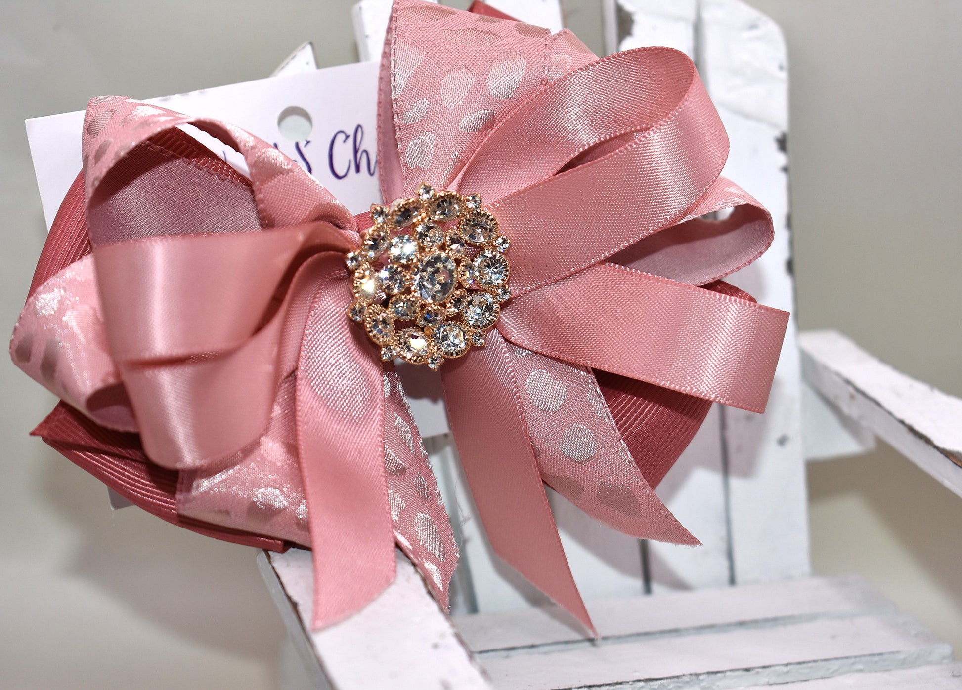 Add a touch of sparkle with this charming bow from Nana's Collection 180. Featuring a soft rose gold pink ribbon topped with a dazzling crystal charm, it’s the perfect playful accent for any outfit. The wrapped alligator clip ensures a secure fit all day long, so you can shine and twirl without a care. Ideal for dressing up your look anytime you want to add a bit of magic!