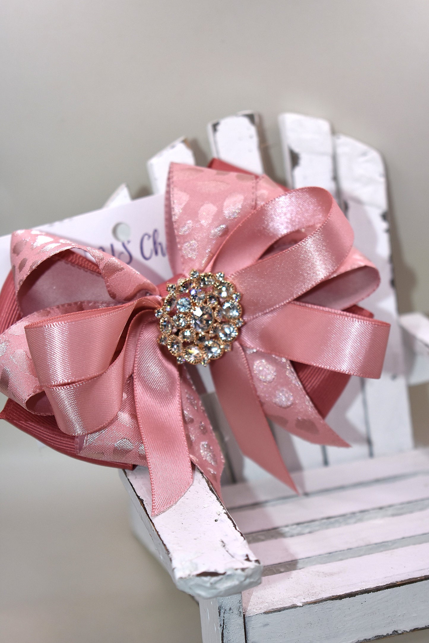 Add a touch of sparkle with this charming bow from Nana's Collection 180. Featuring a soft rose gold pink ribbon topped with a dazzling crystal charm, it’s the perfect playful accent for any outfit. The wrapped alligator clip ensures a secure fit all day long, so you can shine and twirl without a care. Ideal for dressing up your look anytime you want to add a bit of magic!