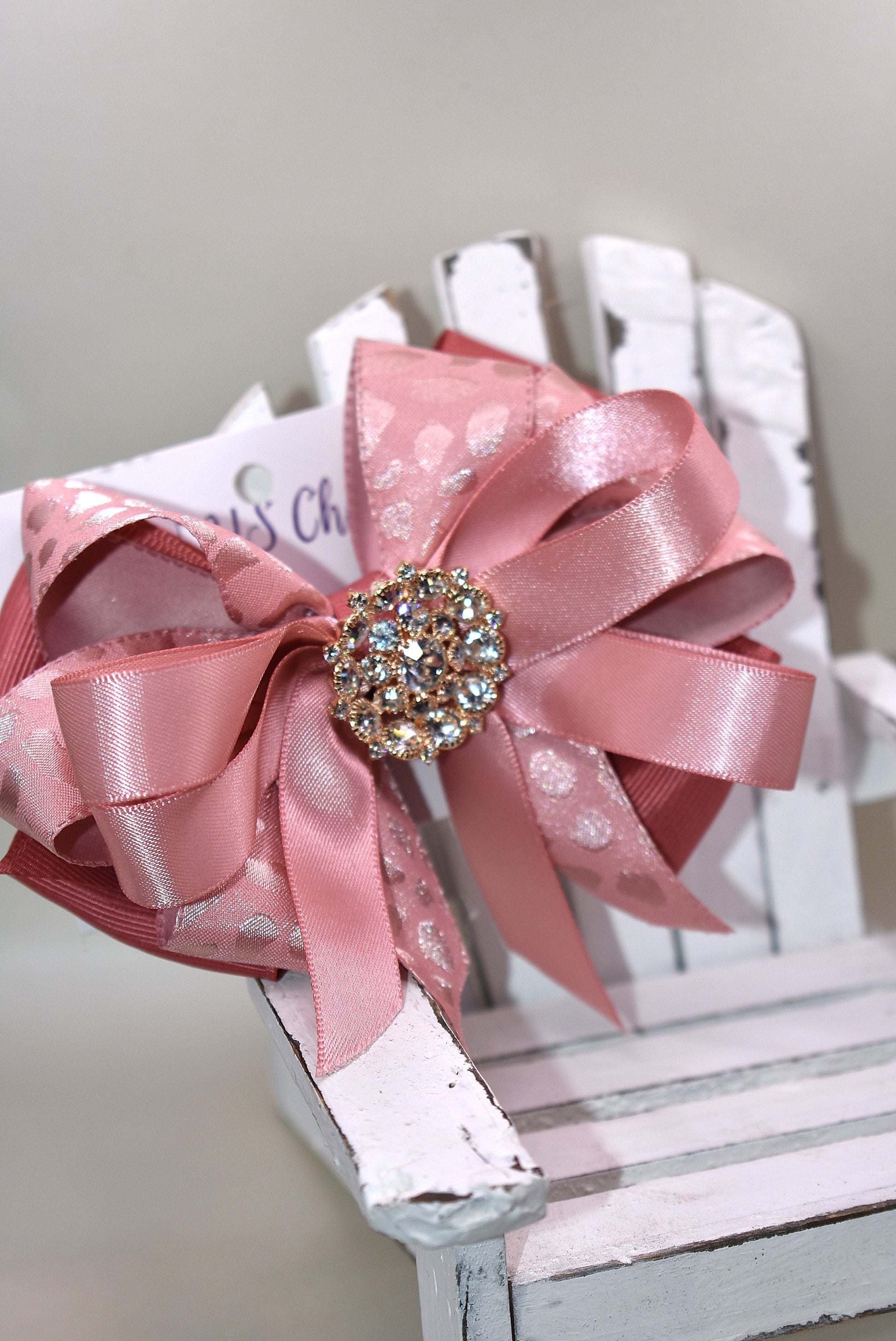 Add a touch of sparkle with this charming bow from Nana's Collection 180. Featuring a soft rose gold pink ribbon topped with a dazzling crystal charm, it’s the perfect playful accent for any outfit. The wrapped alligator clip ensures a secure fit all day long, so you can shine and twirl without a care. Ideal for dressing up your look anytime you want to add a bit of magic!