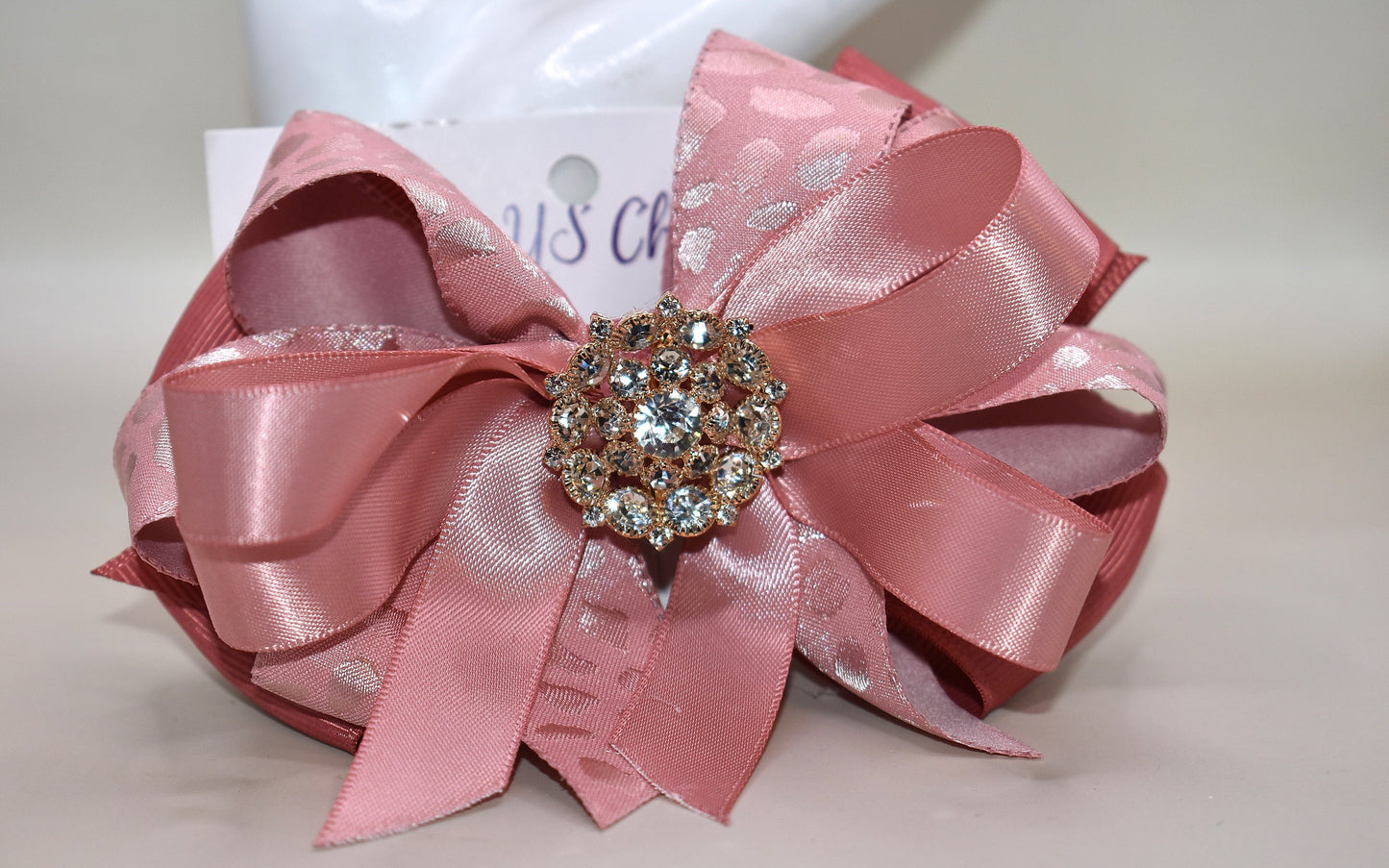Add a touch of sparkle with this charming bow from Nana's Collection 180. Featuring a soft rose gold pink ribbon topped with a dazzling crystal charm, it’s the perfect playful accent for any outfit. The wrapped alligator clip ensures a secure fit all day long, so you can shine and twirl without a care. Ideal for dressing up your look anytime you want to add a bit of magic!