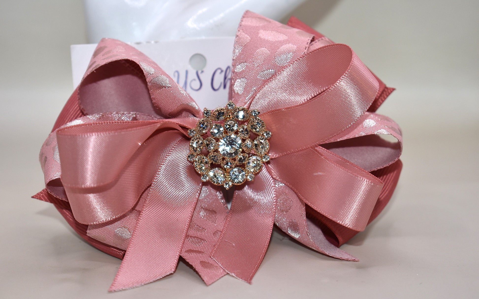 Add a touch of sparkle with this charming bow from Nana's Collection 180. Featuring a soft rose gold pink ribbon topped with a dazzling crystal charm, it’s the perfect playful accent for any outfit. The wrapped alligator clip ensures a secure fit all day long, so you can shine and twirl without a care. Ideal for dressing up your look anytime you want to add a bit of magic!