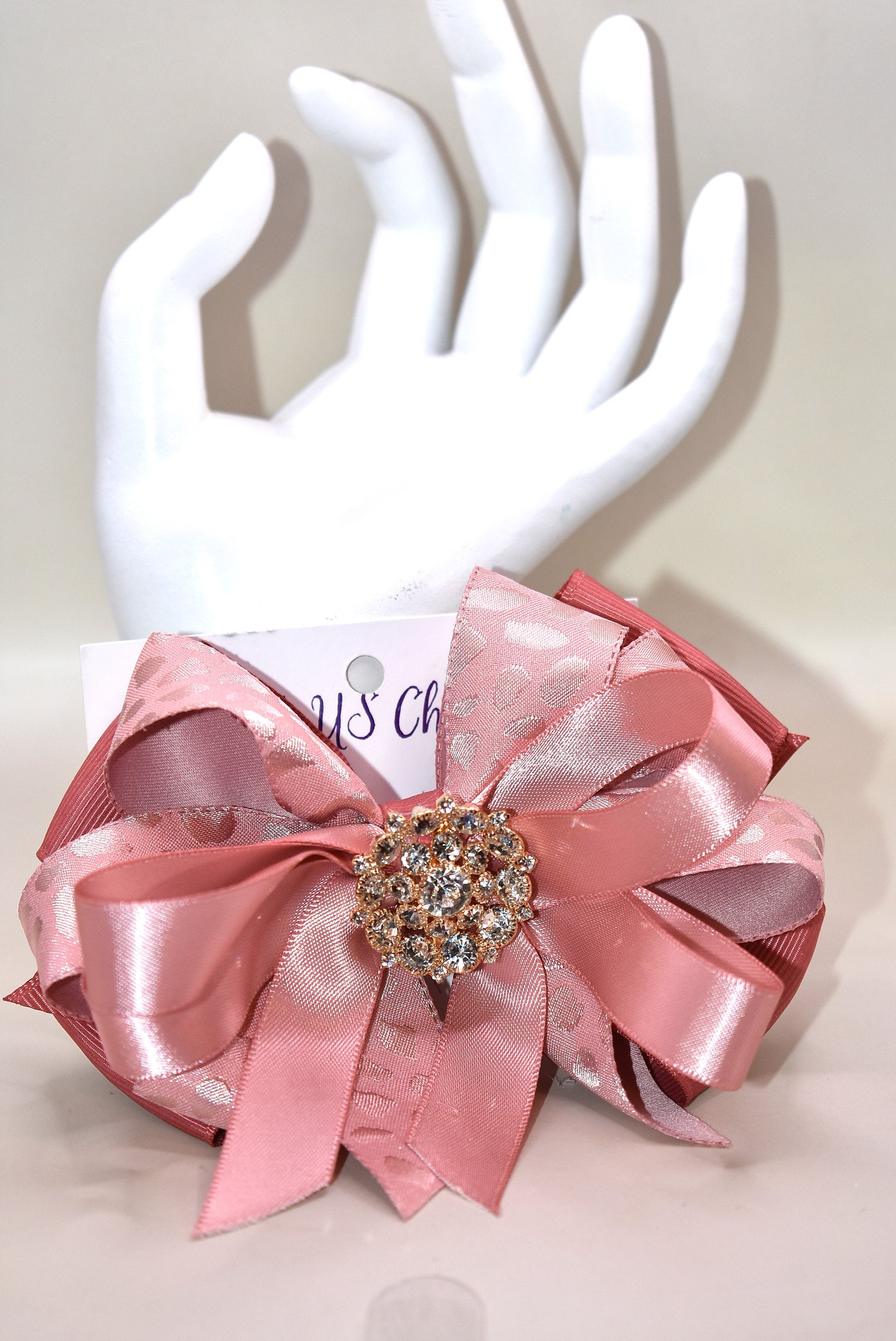 Add a touch of sparkle with this charming bow from Nana's Collection 180. Featuring a soft rose gold pink ribbon topped with a dazzling crystal charm, it’s the perfect playful accent for any outfit. The wrapped alligator clip ensures a secure fit all day long, so you can shine and twirl without a care. Ideal for dressing up your look anytime you want to add a bit of magic!