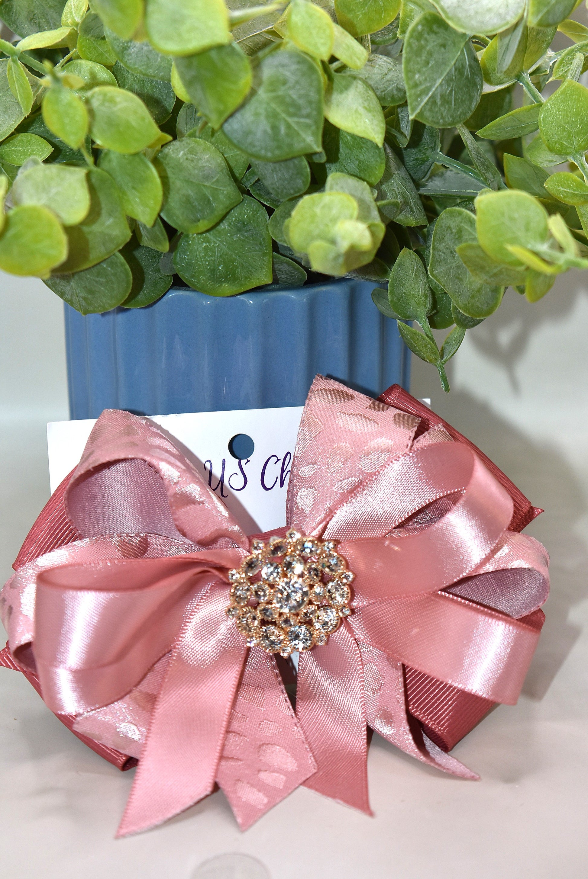 Add a touch of sparkle with this charming bow from Nana's Collection 180. Featuring a soft rose gold pink ribbon topped with a dazzling crystal charm, it’s the perfect playful accent for any outfit. The wrapped alligator clip ensures a secure fit all day long, so you can shine and twirl without a care. Ideal for dressing up your look anytime you want to add a bit of magic!