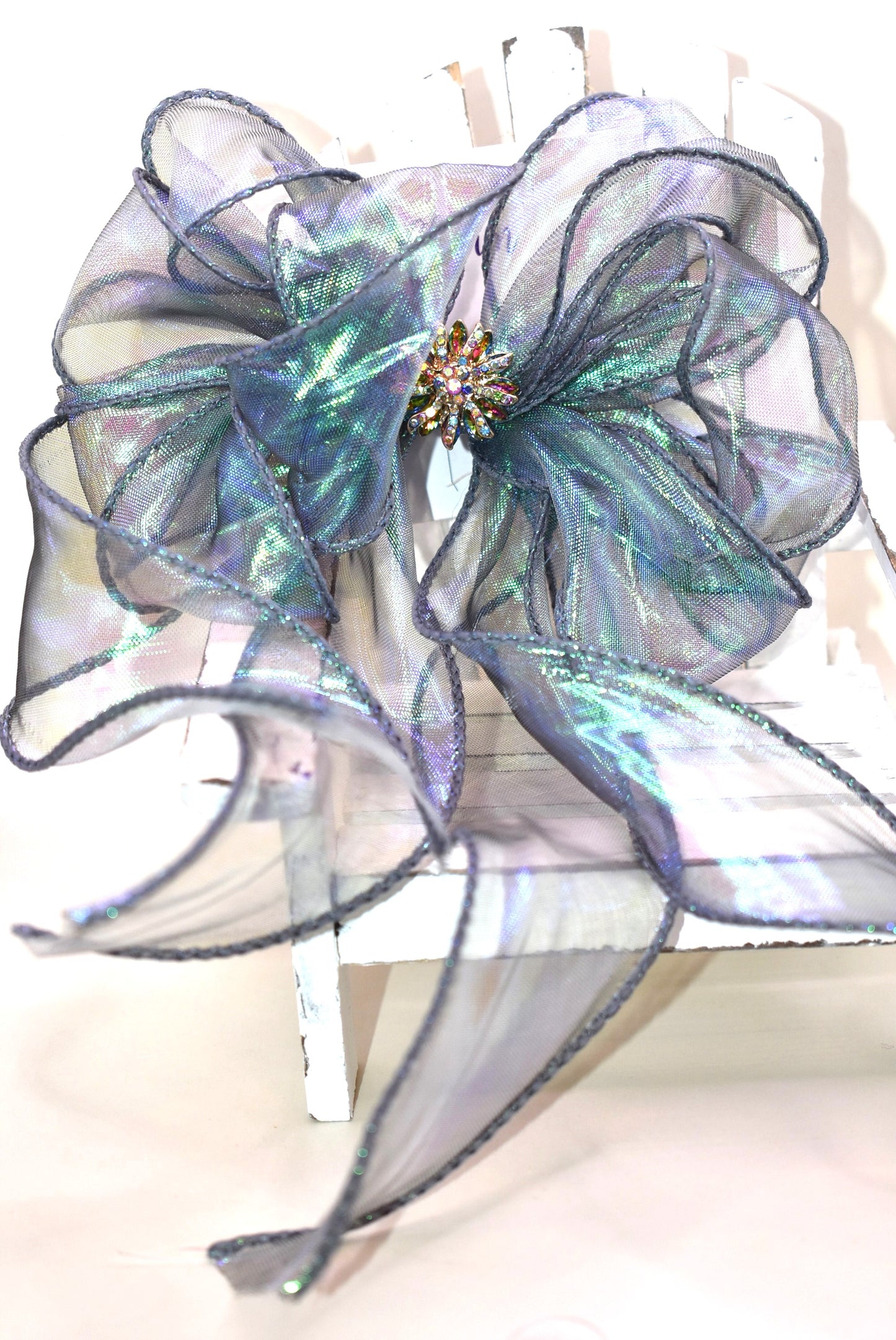 This elegant bow features an iridescent fishtail ribbon in stunning ocean hues, accented by a delicate crystal flower for a refined finish. Its long, flowing design adds graceful movement, while the wrapped alligator clip ensures a secure and comfortable fit. Perfect for adding a touch of sophistication to any look.