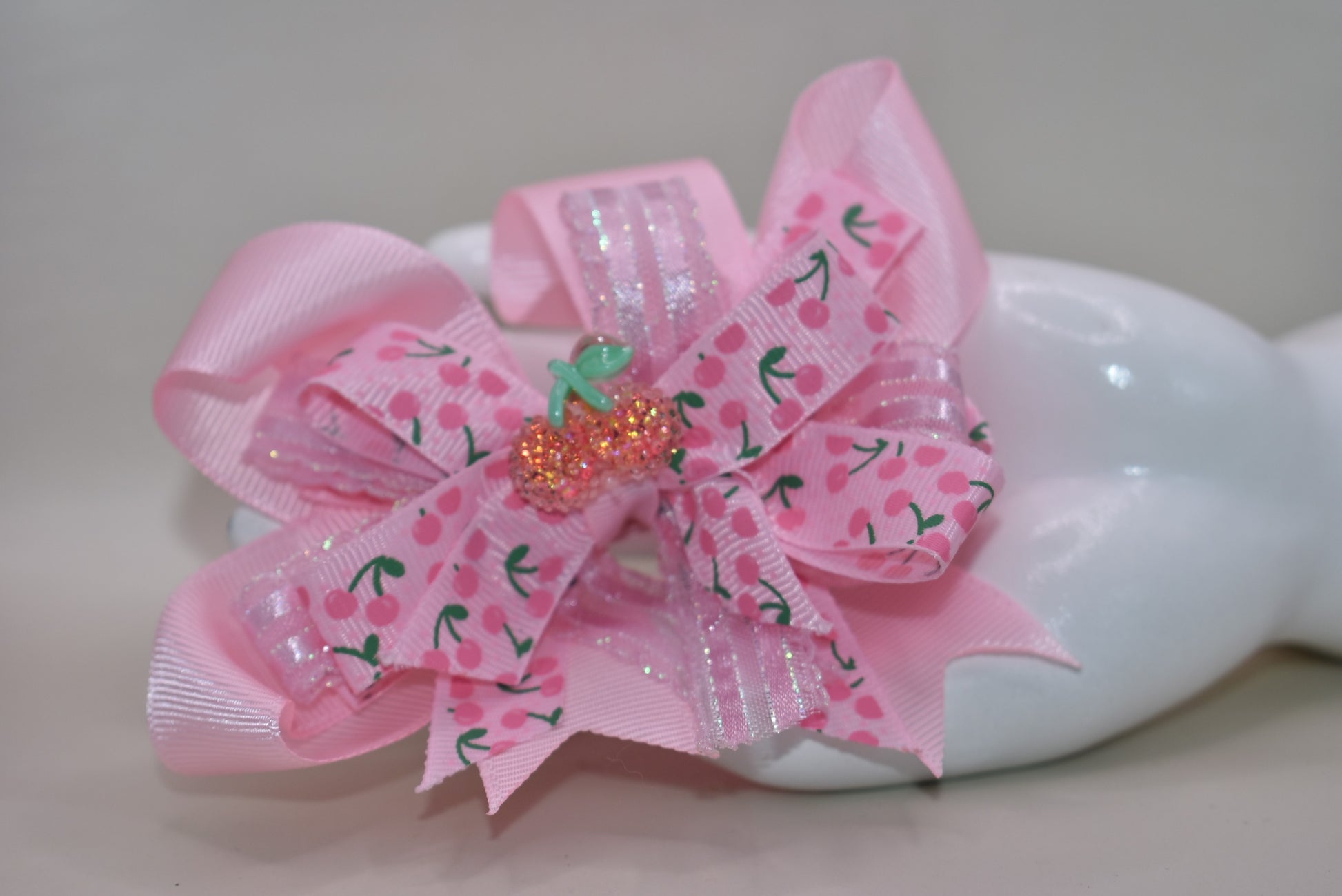 Adorable cherry-themed bow featuring delicate layers of soft pink ribbon that create a charming, dimensional look. The covered alligator clip ensures comfortable wear throughout the day without slipping or pinching. Perfect for adding a sweet pop of personality to any hairstyle, this precious accessory is designed to make you smile every time you catch your reflection. A delightful addition to Nana's Collection that celebrates timeless charm and playful style.