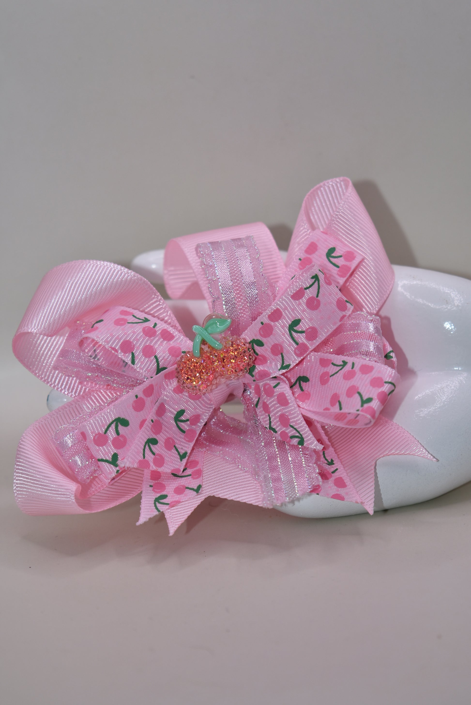 Adorable cherry-themed bow featuring delicate layers of soft pink ribbon that create a charming, dimensional look. The covered alligator clip ensures comfortable wear throughout the day without slipping or pinching. Perfect for adding a sweet pop of personality to any hairstyle, this precious accessory is designed to make you smile every time you catch your reflection. A delightful addition to Nana's Collection that celebrates timeless charm and playful style.