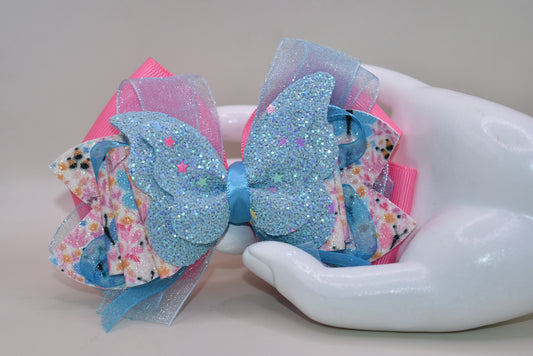 ✨ Introducing a one-of-a-kind bow that's pure magic! 🦋 This stunning creation features luxurious layers of ribbon and faux leather, adorned with delicate butterfly and flower motifs. The showstopper? A gorgeous glitter blue butterfly perched on top that catches the light beautifully. 💙 Secured with a wrapped alligator clip on the back, it's the perfect accessory to add whimsy and charm to any hairstyle. Each piece is uniquely crafted, making it a special treasure from Nana's Collection. 🌸