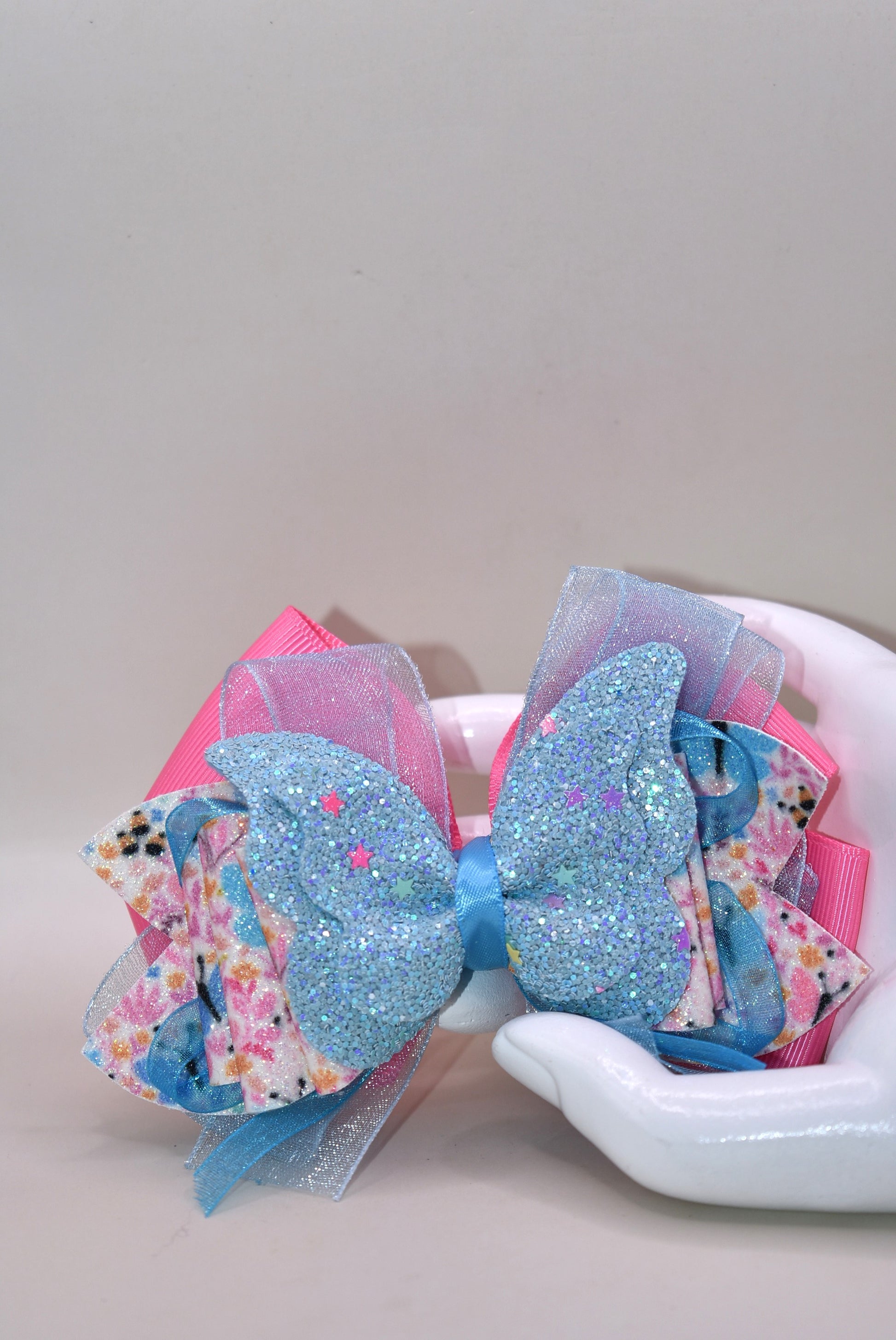 ✨ Introducing a one-of-a-kind bow that's pure magic! 🦋 This stunning creation features luxurious layers of ribbon and faux leather, adorned with delicate butterfly and flower motifs. The showstopper? A gorgeous glitter blue butterfly perched on top that catches the light beautifully. 💙 Secured with a wrapped alligator clip on the back, it's the perfect accessory to add whimsy and charm to any hairstyle. Each piece is uniquely crafted, making it a special treasure from Nana's Collection. 🌸