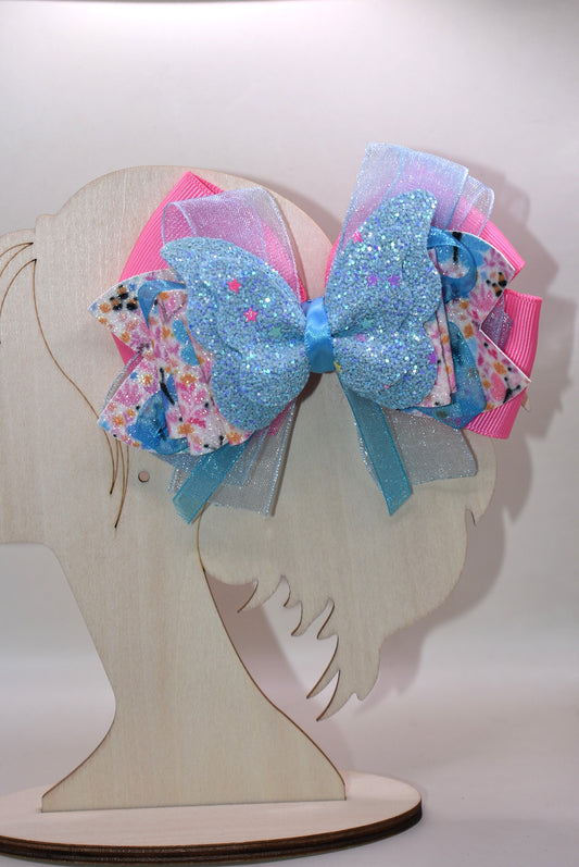 ✨ Introducing a one-of-a-kind bow that's pure magic! 🦋 This stunning creation features luxurious layers of ribbon and faux leather, adorned with delicate butterfly and flower motifs. The showstopper? A gorgeous glitter blue butterfly perched on top that catches the light beautifully. 💙 Secured with a wrapped alligator clip on the back, it's the perfect accessory to add whimsy and charm to any hairstyle. Each piece is uniquely crafted, making it a special treasure from Nana's Collection. 🌸